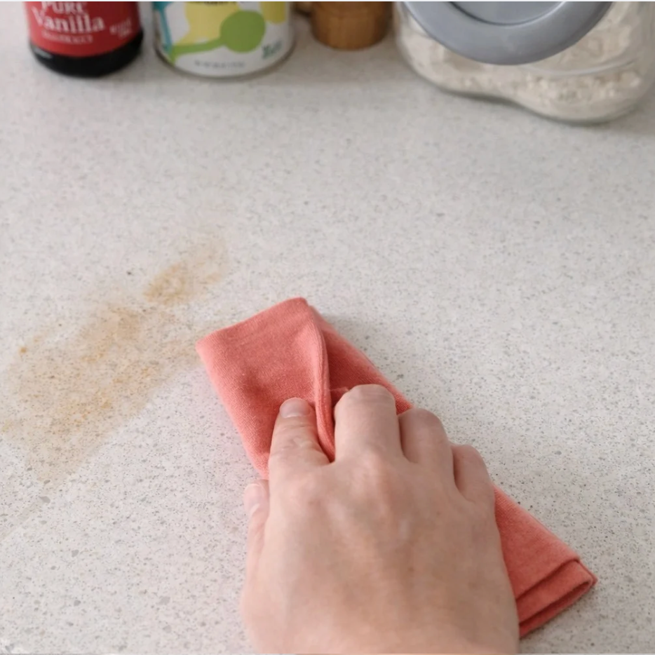 Hand using a pink cloth to wipe a stain on a white countertop with various items in the background.